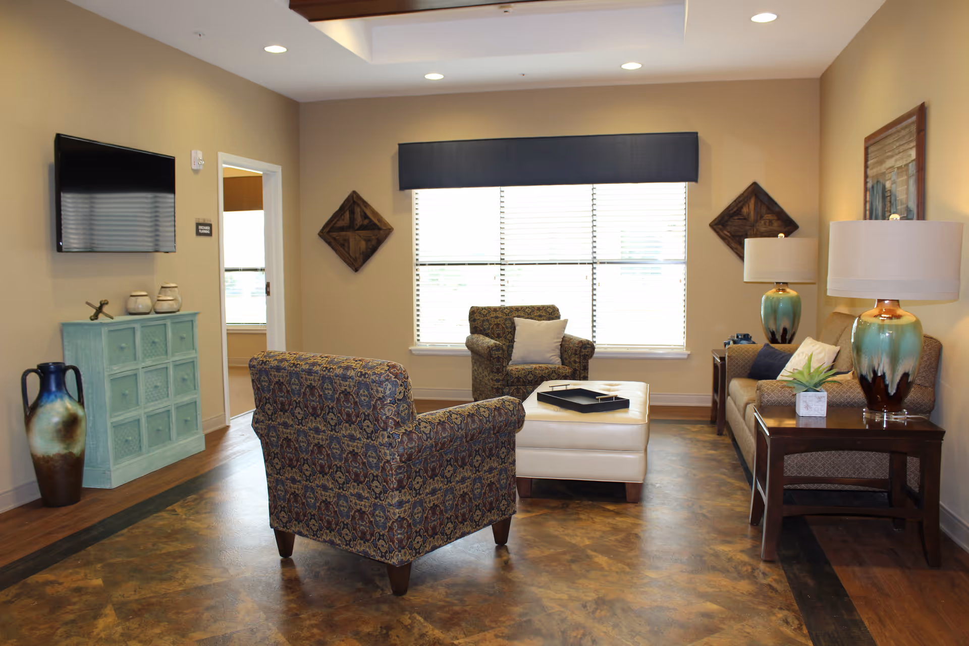 A cozy living room area with two patterned armchairs, a beige ottoman, and a sofa with decorative pillows. There are two matching table lamps with green and brown bases on wooden side tables. A large window with blinds and a dark valance lets in natural light. On the left wall, a flat-screen TV is mounted above a light blue cabinet with decorative items. The floor has a brown and tan patterned surface, and the walls are painted beige with some wall art.