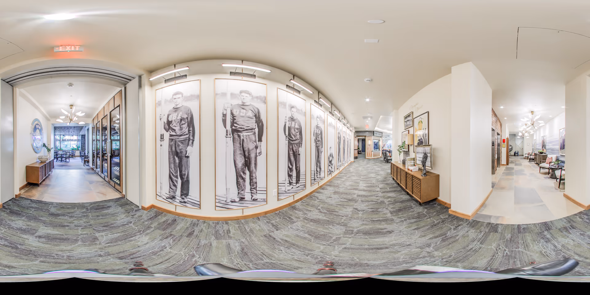 A wide hallway in a senior living facility with a carpeted floor and white walls. One side of the hallway features large black and white portraits of athletes wearing USA sweaters. The opposite side has a wooden console table with decorative items and framed pictures above it. At the end of the hallway, there is a seating area with chairs and tables near large windows letting in natural light. The ceiling has recessed lighting and modern light fixtures.
