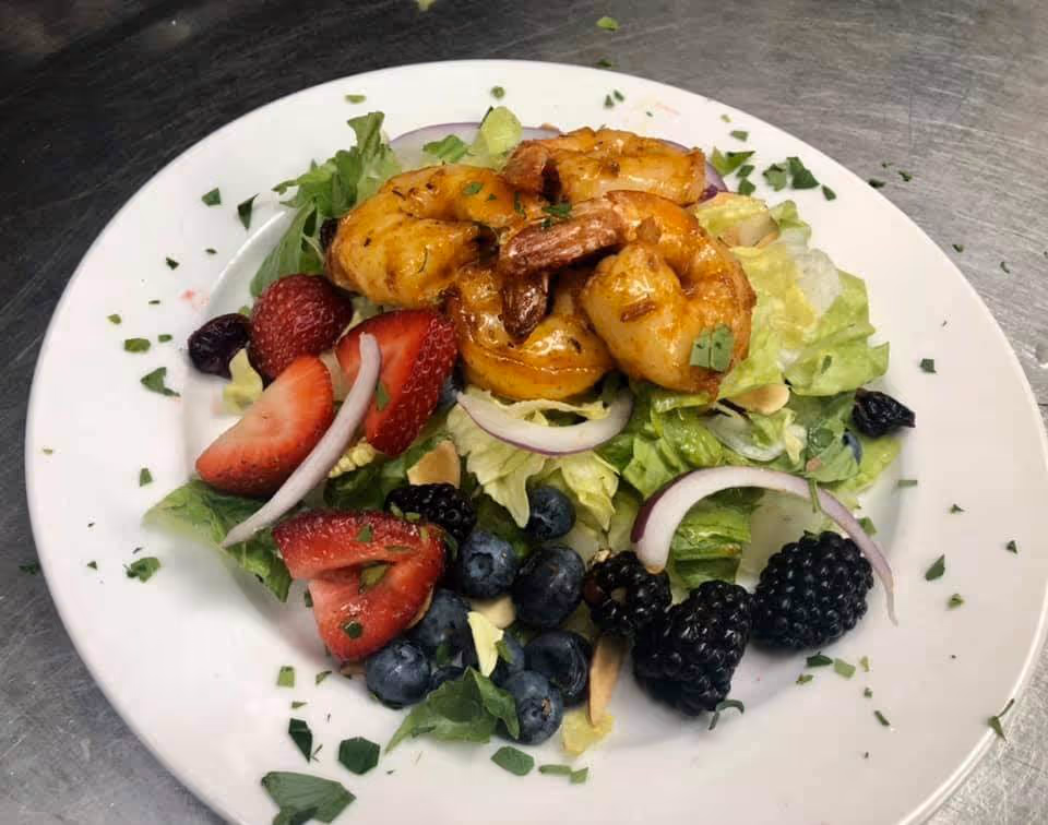 Grilled shrimp served on a mixed green salad topped with strawberries, blueberries, blackberries, and red onion on a white plate.