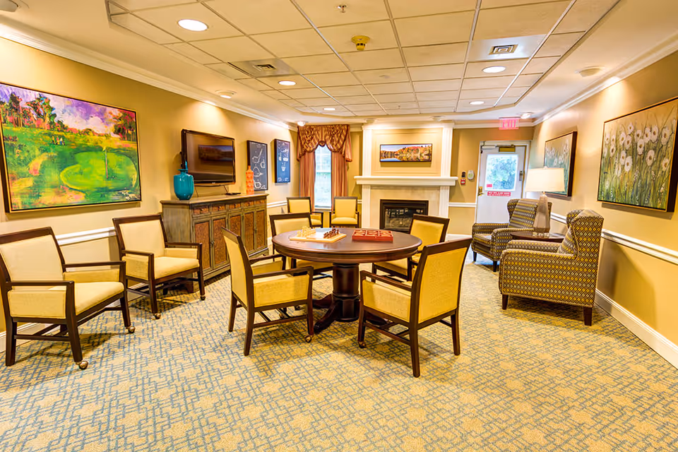 Bright furnished common lounge with a round table surrounded by chairs, a fireplace, TV, and wall artwork.