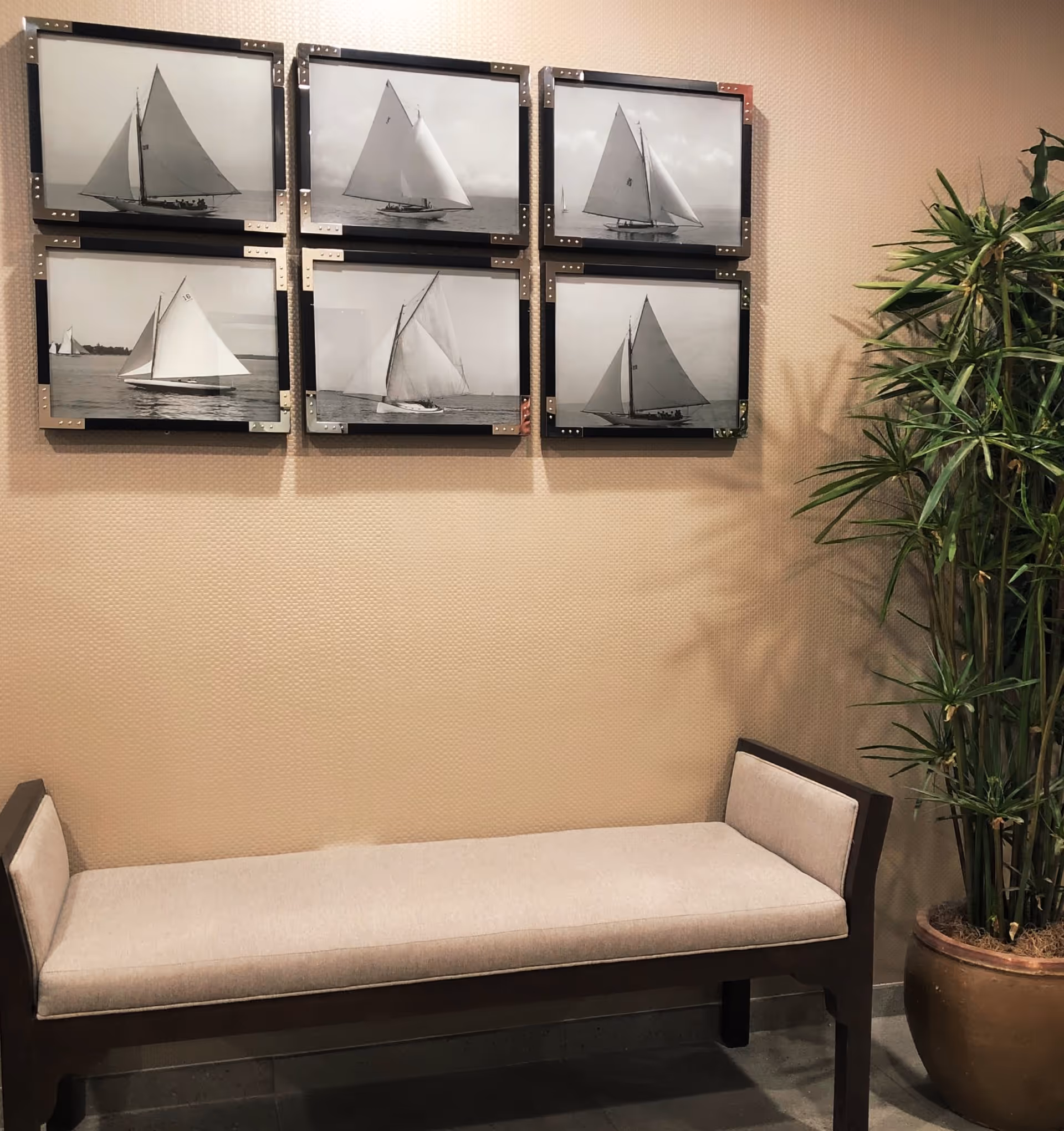 A beige cushioned bench with dark wooden frame is placed against a textured beige wall. Above the bench, there are six black and white framed photographs of sailboats arranged in two rows of three. To the right of the bench, there is a tall potted plant with long green leaves.