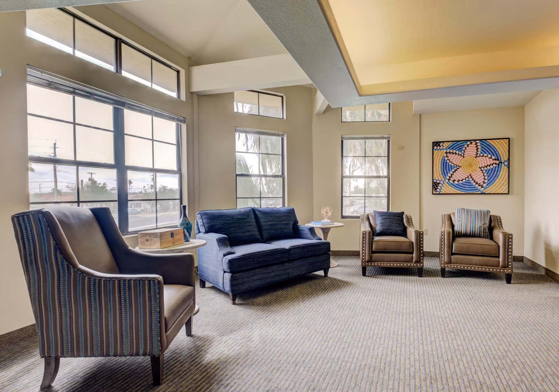 A bright and spacious living room area with large windows letting in natural light. The room features a blue loveseat, two brown armchairs with striped cushions, and a striped armchair. There are two small round side tables, one with a decorative blue vase and the other with a small sculpture and books. A colorful framed artwork hangs on the wall.