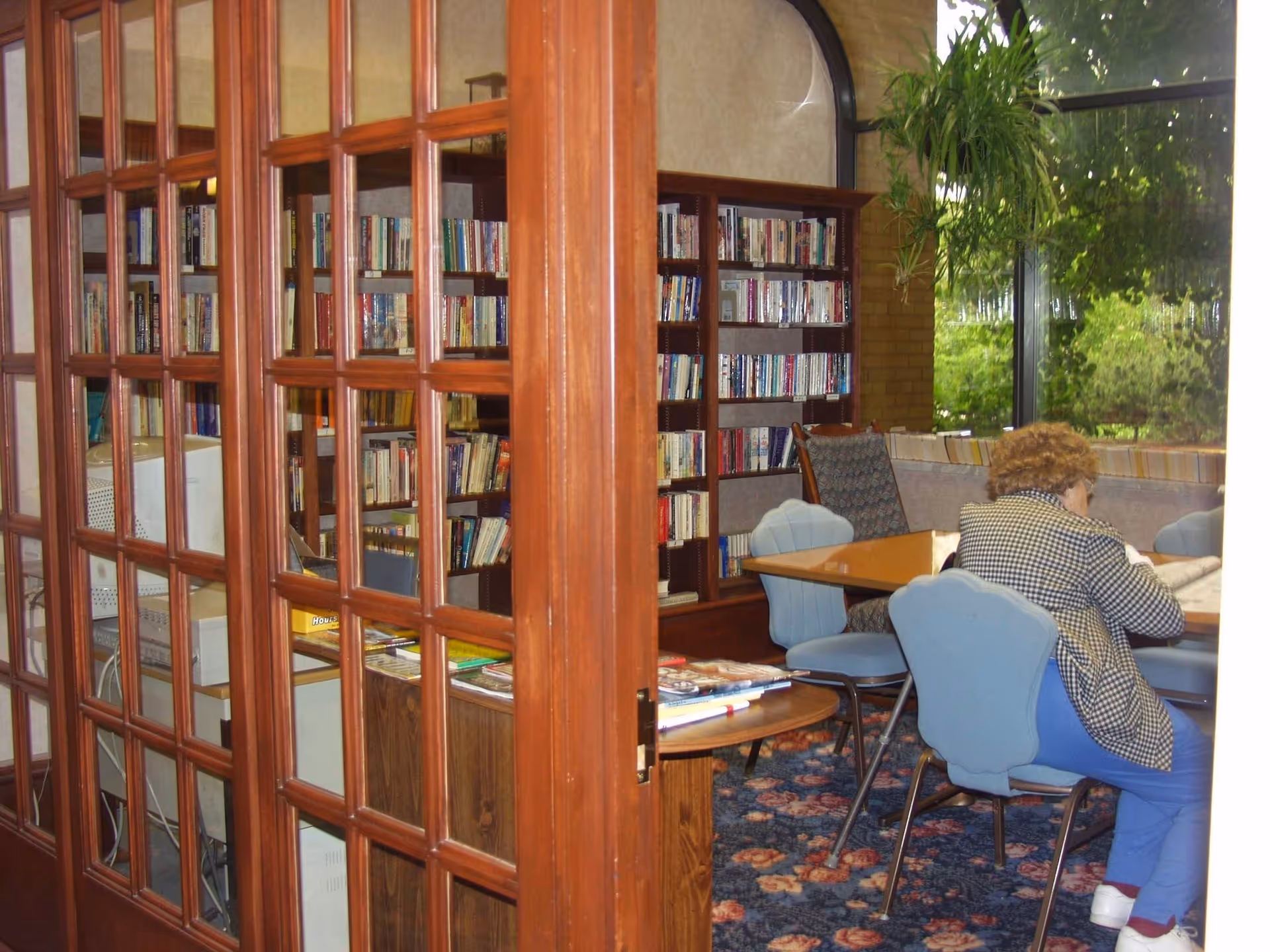 A communal library/reading room with bookshelves, a table and chairs, and a person seated reading near a window.