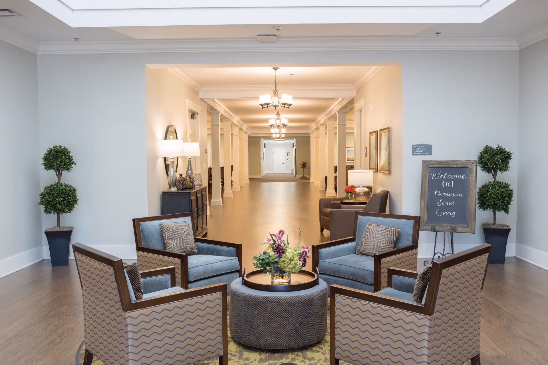 A bright senior living lobby with a circular seating area of armchairs around a central ottoman and a long hallway beyond.