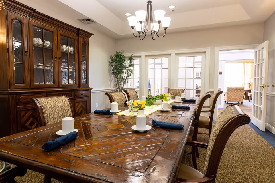 A dining room with a large wooden table set with white cups on saucers and dark blue napkins. The table has a yellow and white floral centerpiece. There are upholstered chairs around the table, a wooden cabinet with glass doors displaying china, and a potted plant in the corner. French doors lead to another room with seating visible.