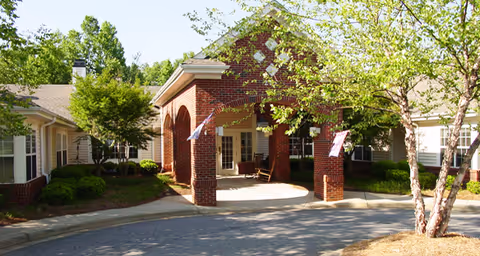 Covered brick entrance with a porte-cochère, flags, and landscaped driveway in front of a senior living building.