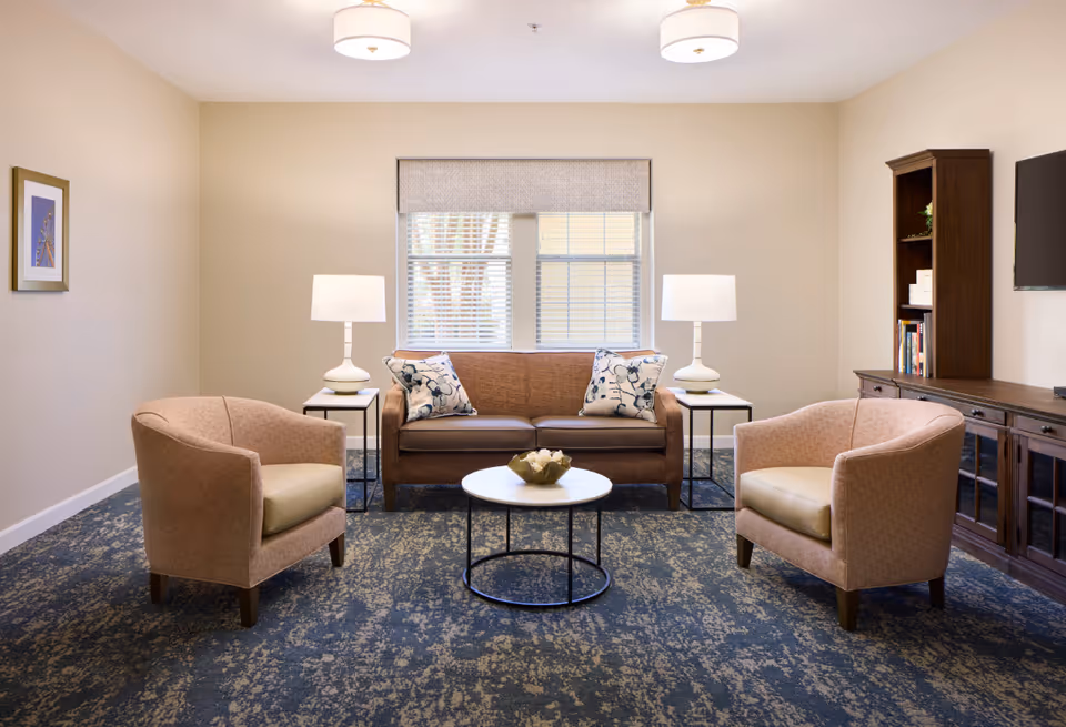 A cozy living room with a brown sofa adorned with two patterned pillows, flanked by two white table lamps on side tables. Two beige armchairs face the sofa across a round coffee table with a decorative centerpiece. A wooden cabinet with books and decor is against the right wall, and a flat-screen TV is mounted above it. A window with blinds and a valance is centered on the back wall, and a framed picture hangs on the left wall. The floor is covered with a blue patterned carpet.