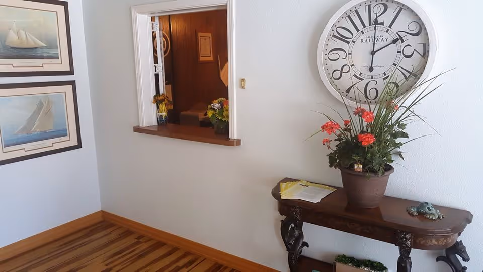 Interior corner of a room with light-colored walls and wooden floor. On the left wall, there are two framed pictures of sailboats. A small pass-through window with a wooden ledge opens into another room with wood-paneled walls and a small vase of flowers on the ledge. On the right wall, a large round clock with black numbers hangs above a dark wooden console table. The table holds a potted plant with red flowers, some papers, and a small decorative frog figurine.