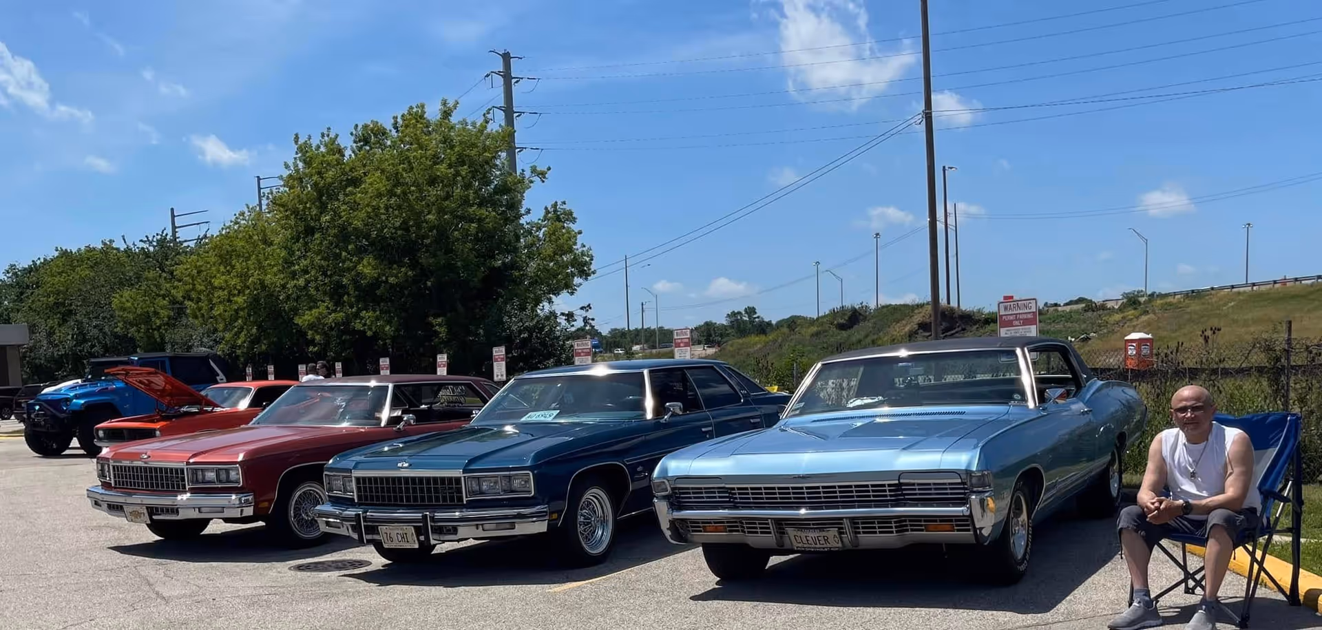 Three classic cars parked side by side in a parking lot on a sunny day with a man sitting on a folding chair next to the light blue car. Trees and a fence are visible in the background under a blue sky with some clouds.