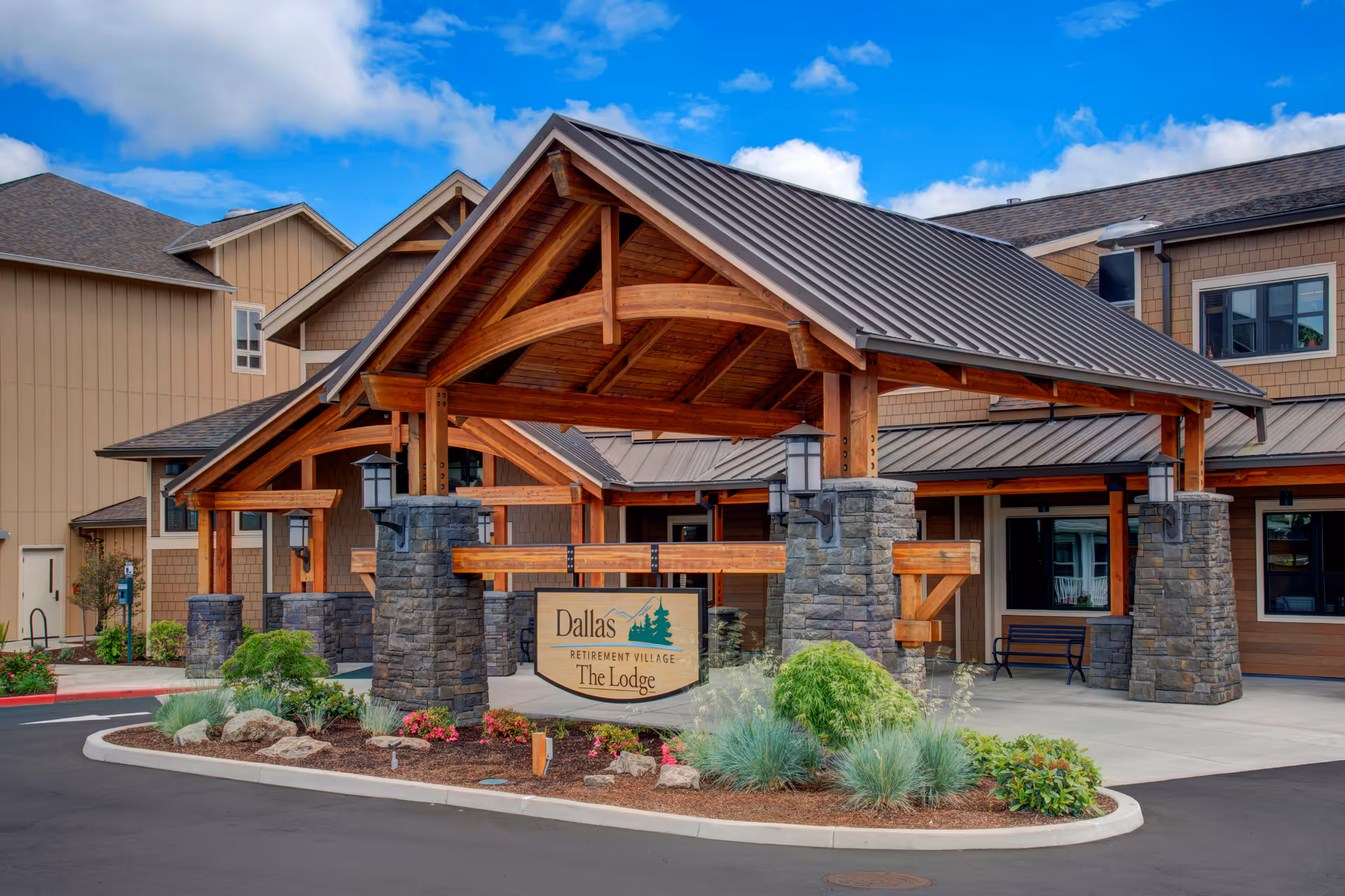 Exterior view of the entrance to Dallas Retirement Village The Lodge, featuring a wooden and stone covered driveway with landscaped plants and a clear blue sky.