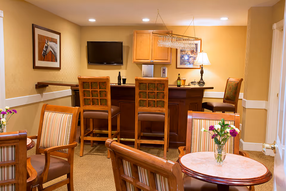 Cozy common room featuring a wooden bar with high stools, round tables with chairs, and a wall-mounted TV.