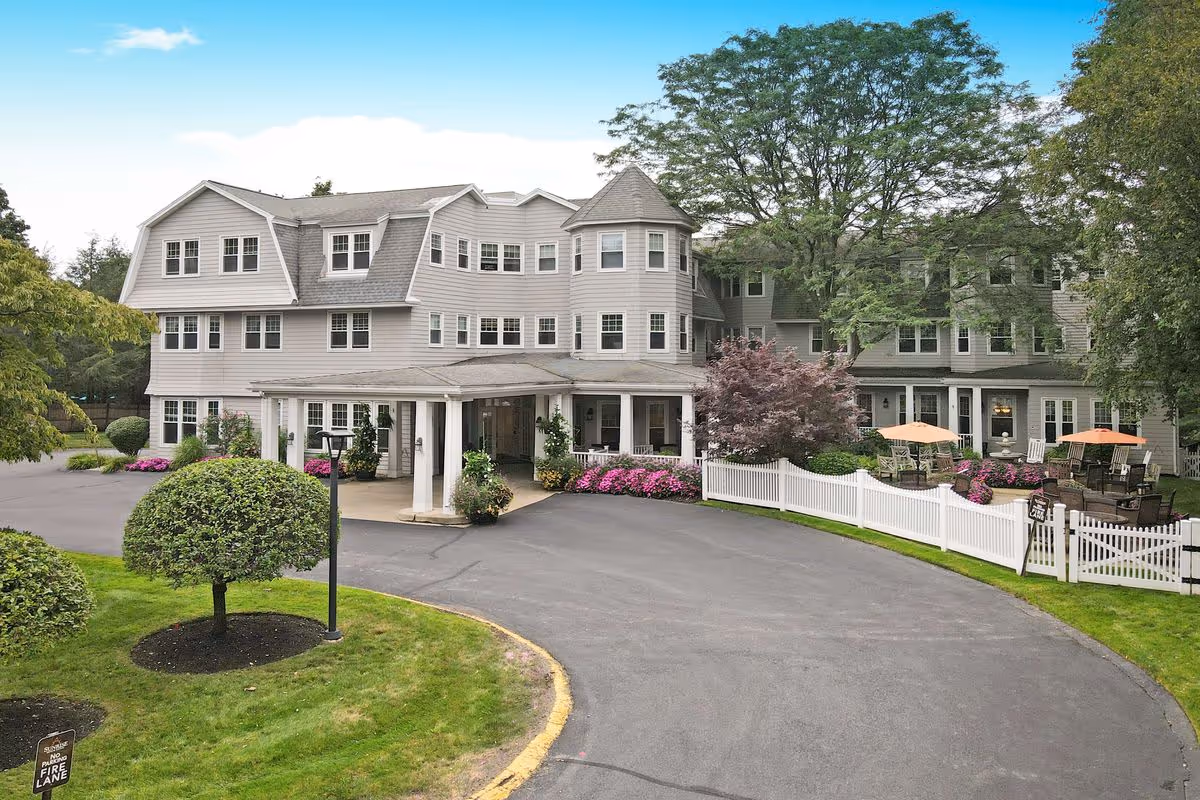 Exterior view of a large, multi-story senior living facility with gray siding, numerous windows, and a covered entrance. The driveway curves in front of the building, surrounded by well-maintained landscaping including trimmed bushes, flowering plants, and trees. There is a white picket fence enclosing an outdoor patio area with tables, chairs, and umbrellas.
