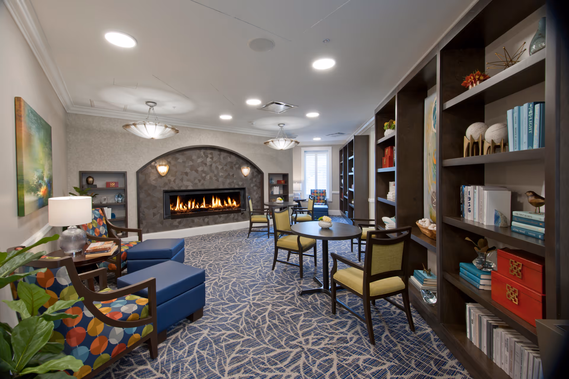 A cozy senior living common area with a modern fireplace set into a textured wall. The room features patterned carpet, several round tables with chairs, colorful armchairs with matching ottomans, bookshelves filled with books and decorative items, and soft lighting from ceiling fixtures and table lamps.
