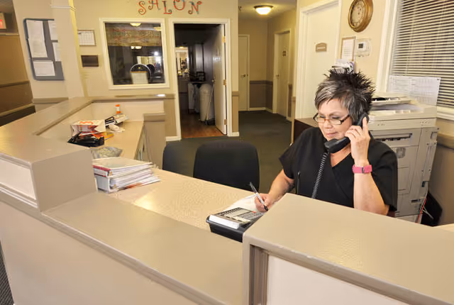 A woman with short hair and glasses is sitting at a reception desk, talking on the phone and writing on a notepad. The desk has office supplies and papers, and behind her is a hallway with a door labeled 'Salon' and other rooms.