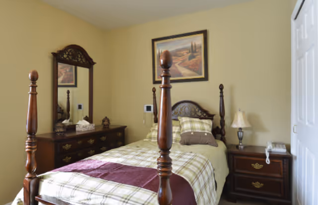 A traditional bedroom with a four-poster bed, plaid bedding, a wooden dresser with mirror, and a nightstand with lamp.