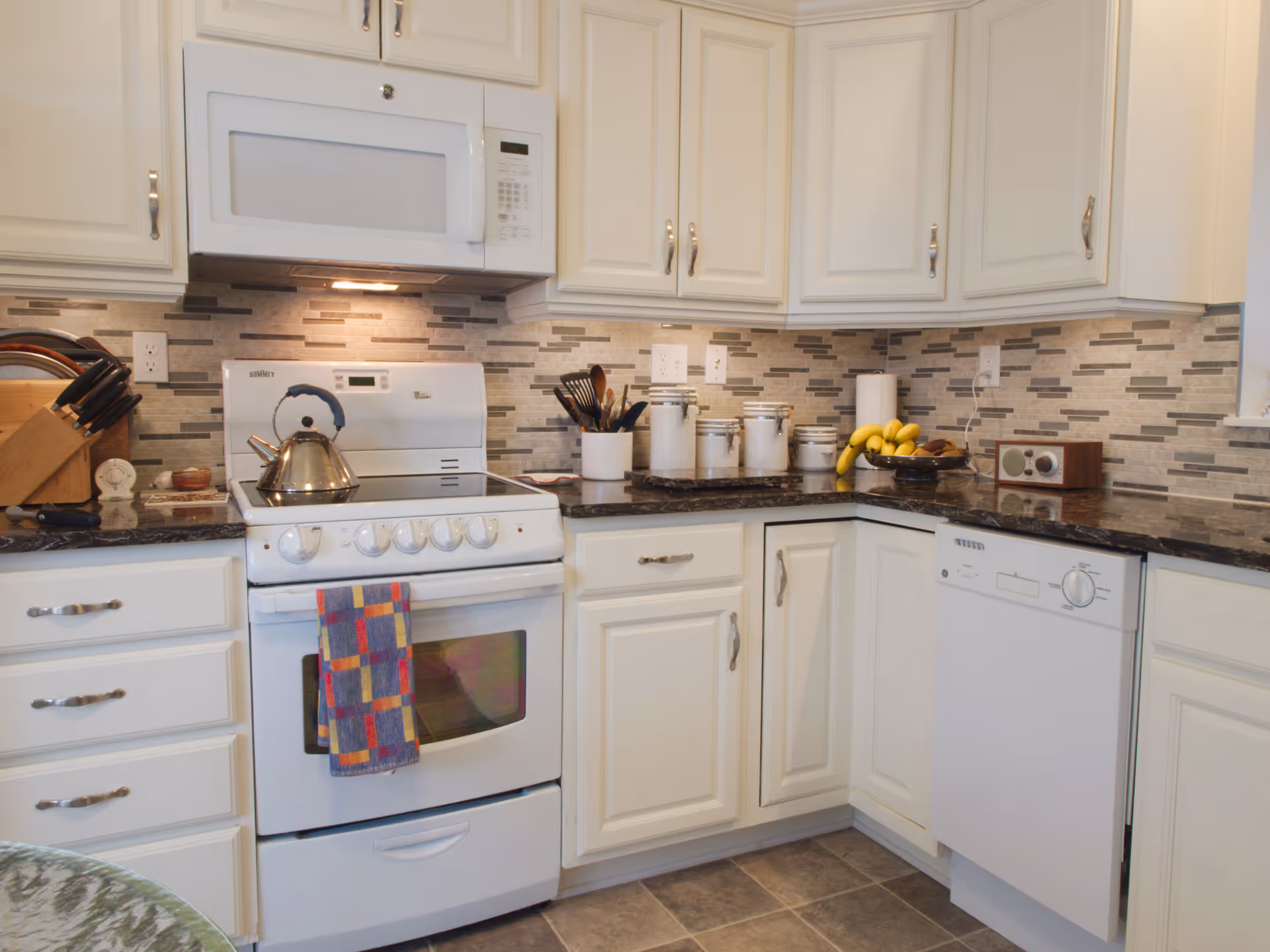 A clean and modern kitchen with white cabinets, a white stove with a colorful towel hanging on the handle, a stainless steel kettle on the stove, a white microwave above the stove, a black granite countertop with various kitchen utensils, white canisters, a bowl of bananas, a paper towel holder, and a white dishwasher. The backsplash features a mosaic tile design in neutral tones.