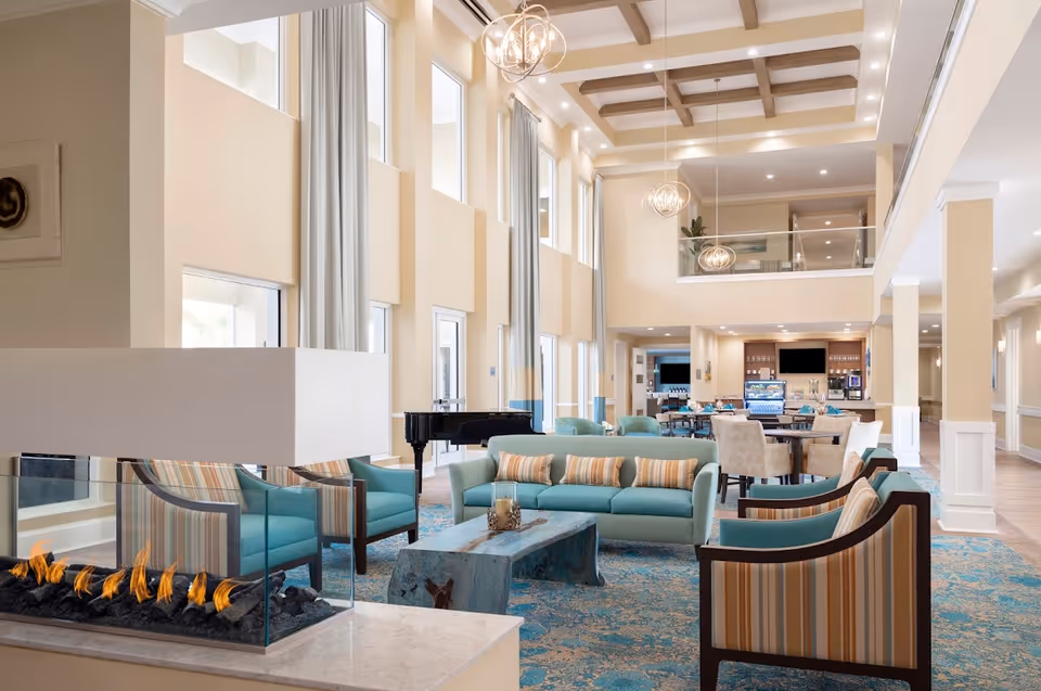 Spacious senior living common area with high ceilings, multiple seating groups, a glass-front fireplace, dining tables and a piano.