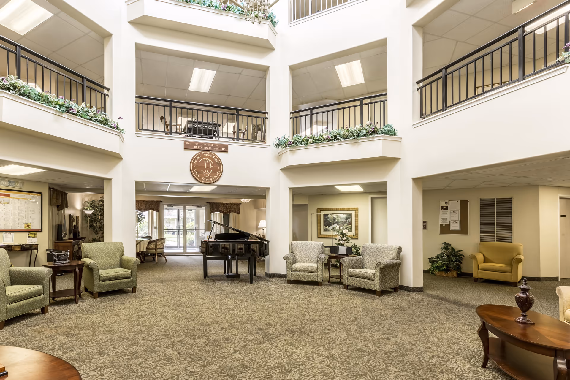 A spacious lobby area featuring green armchairs, a grand piano, and decorative plants. The high ceilings and open layout create a welcoming atmosphere, with natural light coming from large windows.