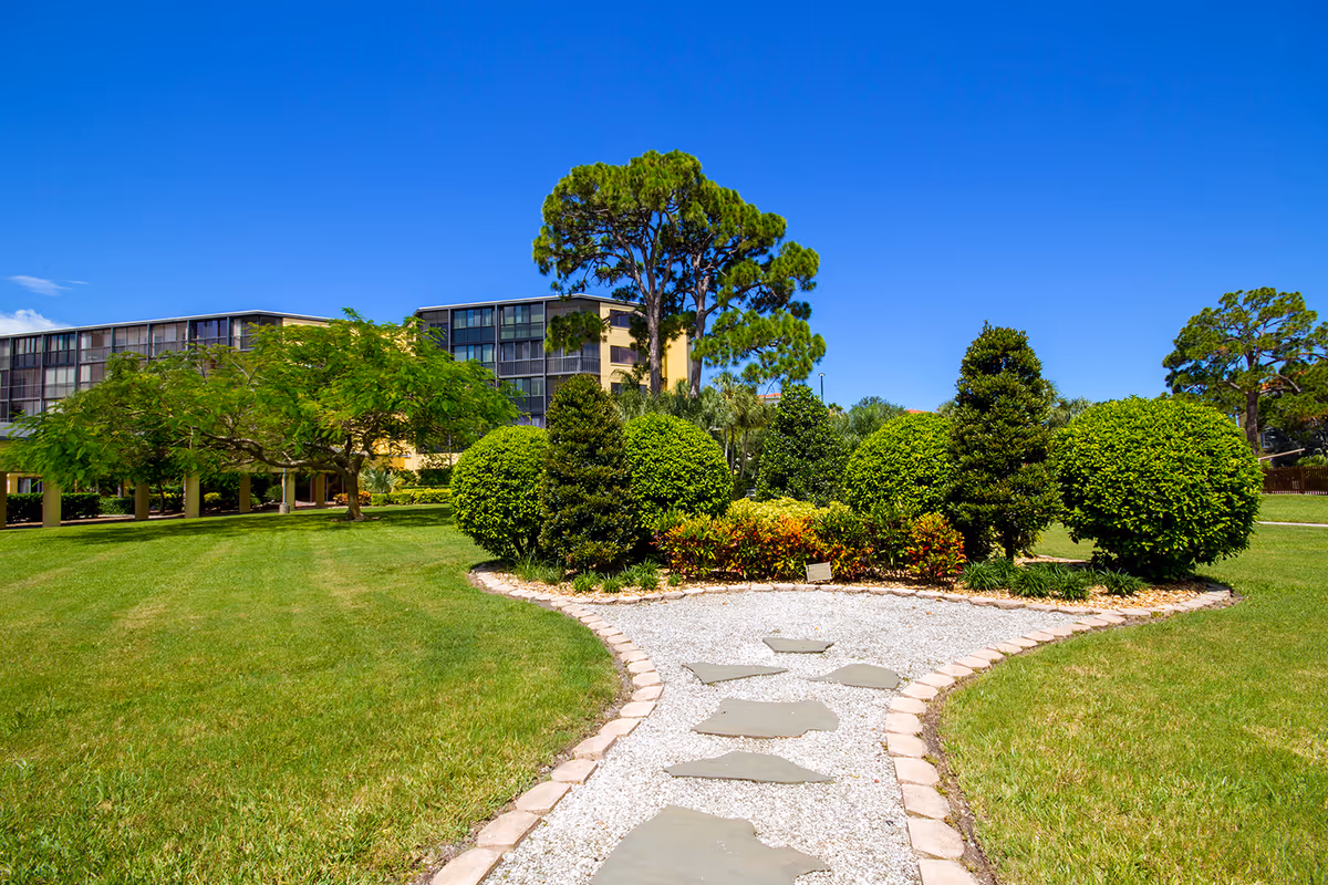 A landscaped outdoor garden area with a gravel and stone pathway leading to a variety of neatly trimmed bushes and trees. In the background, there is a multi-story building under a clear blue sky.