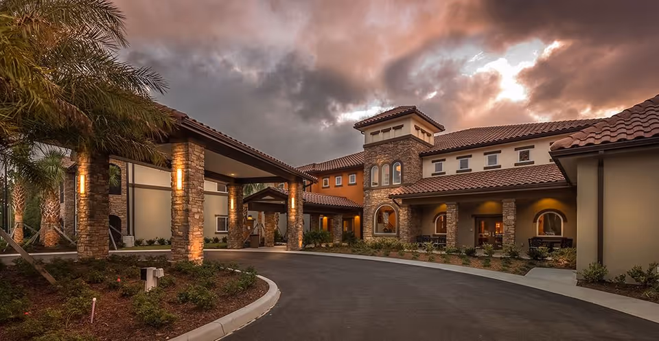 Exterior view of HarborChase of Palm Coast facility at dusk, featuring a curved driveway, stone pillars, warm lighting, and a tiled roof under a cloudy sky.