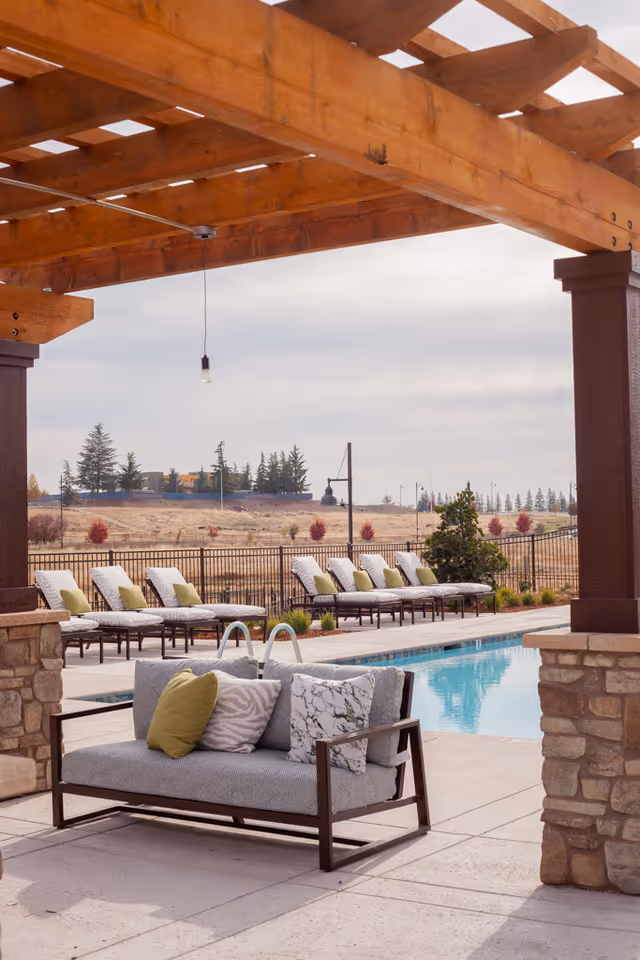 Outdoor pool area with a wooden pergola overhead, a cushioned bench with decorative pillows in the foreground, and a row of lounge chairs with green pillows lined up along the poolside. The background shows a fenced area with trees and a cloudy sky.