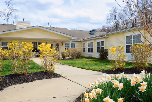 Exterior view of a single-story yellow building with white trim, surrounded by green grass, yellow flowering bushes, and white and orange daffodils along a concrete walkway under a partly cloudy sky.