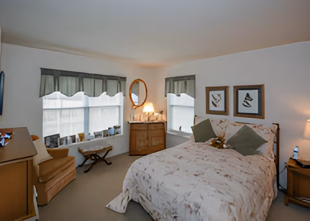 A cozy bedroom with a large bed covered in a light-colored patterned comforter and two green pillows. There are two framed artworks above the bed. The room has two windows with green valances, a wooden dresser with a round mirror and a lamp on top, a beige armchair, and a small wooden stool. The walls are white and the carpet is light brown.
