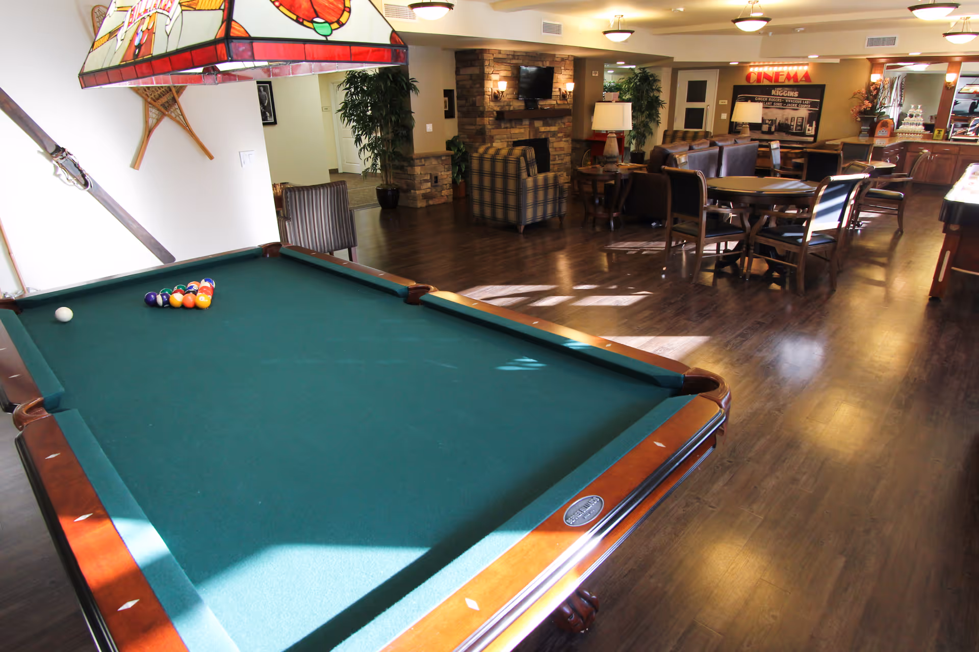 Interior view of a recreational room featuring a pool table with balls arranged for a game, a hanging stained glass light fixture above the table, several chairs and tables, a stone fireplace with a mounted TV, and a sign that reads 'CINEMA' in the background.