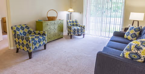 A bright living room with beige carpet, featuring a dark blue sofa with patterned cushions, two patterned armchairs, a green decorative cabinet with a basket on top, a floor lamp, and large vertical blinds covering a sliding glass door that leads outside.