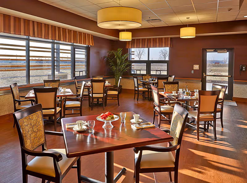 A bright dining room with several wooden tables and chairs arranged neatly. Each table is set with cups, glasses, and napkins. Large windows allow natural light to fill the room, and there are three large ceiling lights. A potted plant is visible in the corner near the windows, and a door leads outside.