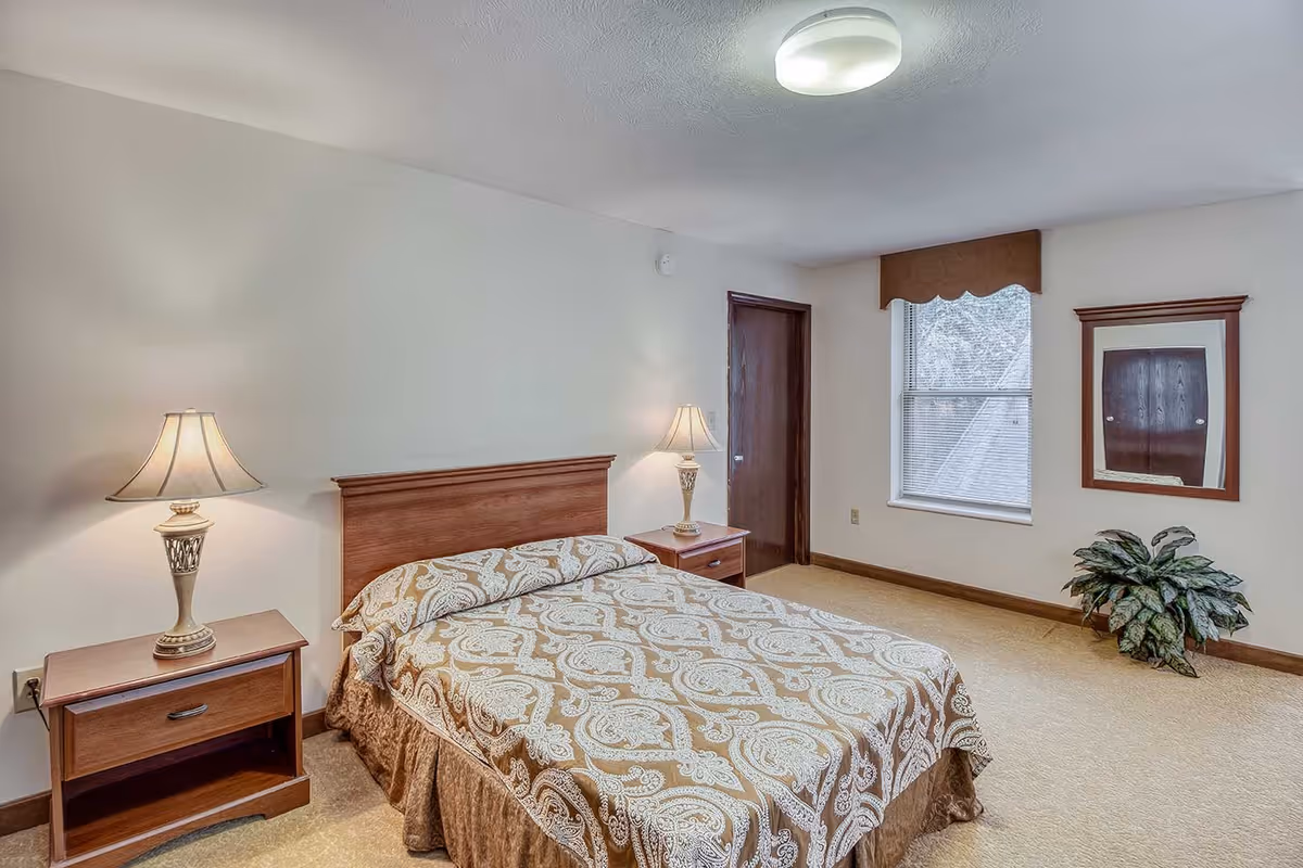A bedroom with a double bed covered in a patterned beige and white bedspread. There are two wooden nightstands on either side of the bed, each with a lamp. The room has beige carpet, a window with a brown valance, a wooden door, a framed mirror on the wall, and a potted plant in the corner.