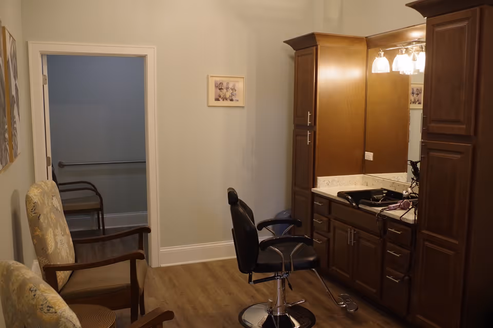 Interior room with a salon chair in front of a large mirror and wooden cabinets. Two upholstered armchairs are positioned along the left wall. The room has light-colored walls and wood flooring, with a doorway leading to another room with a handrail visible.