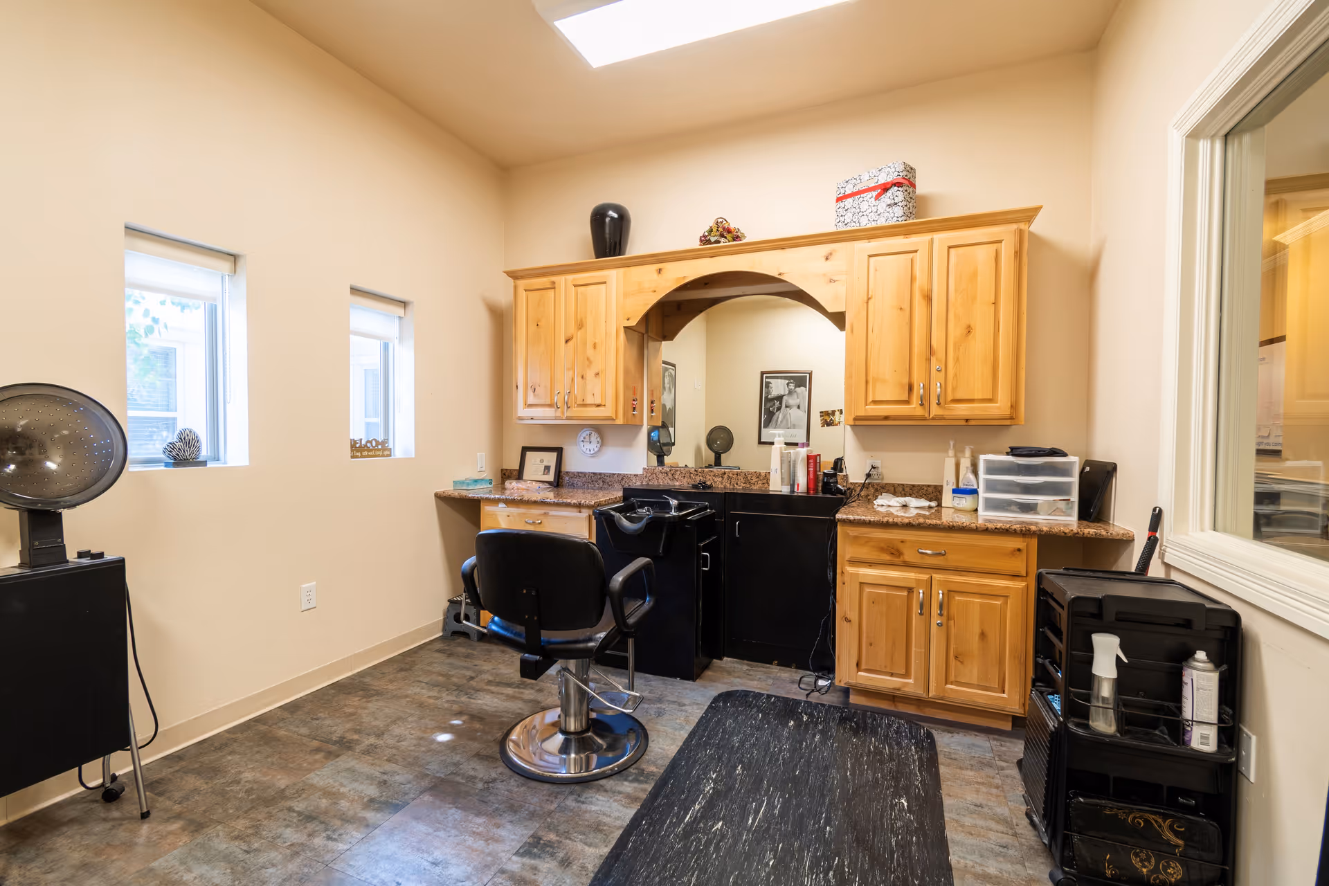 Interior view of a hair salon area within a senior living facility, featuring a black salon chair in front of a counter with a large mirror, wooden cabinets above and below the counter, and various hair care products. There is a hair dryer on a stand to the left and a rolling cart with supplies to the right. The room has beige walls, two small windows, and a skylight.