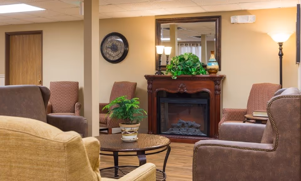 A cozy living room area in an assisted living facility featuring a wooden electric fireplace with decorative plants and a mirror above it. Surrounding the fireplace are several upholstered armchairs in brown and beige tones, a round coffee table with a potted plant, a standing lamp, and a wall clock. The room has warm beige walls and wooden flooring.