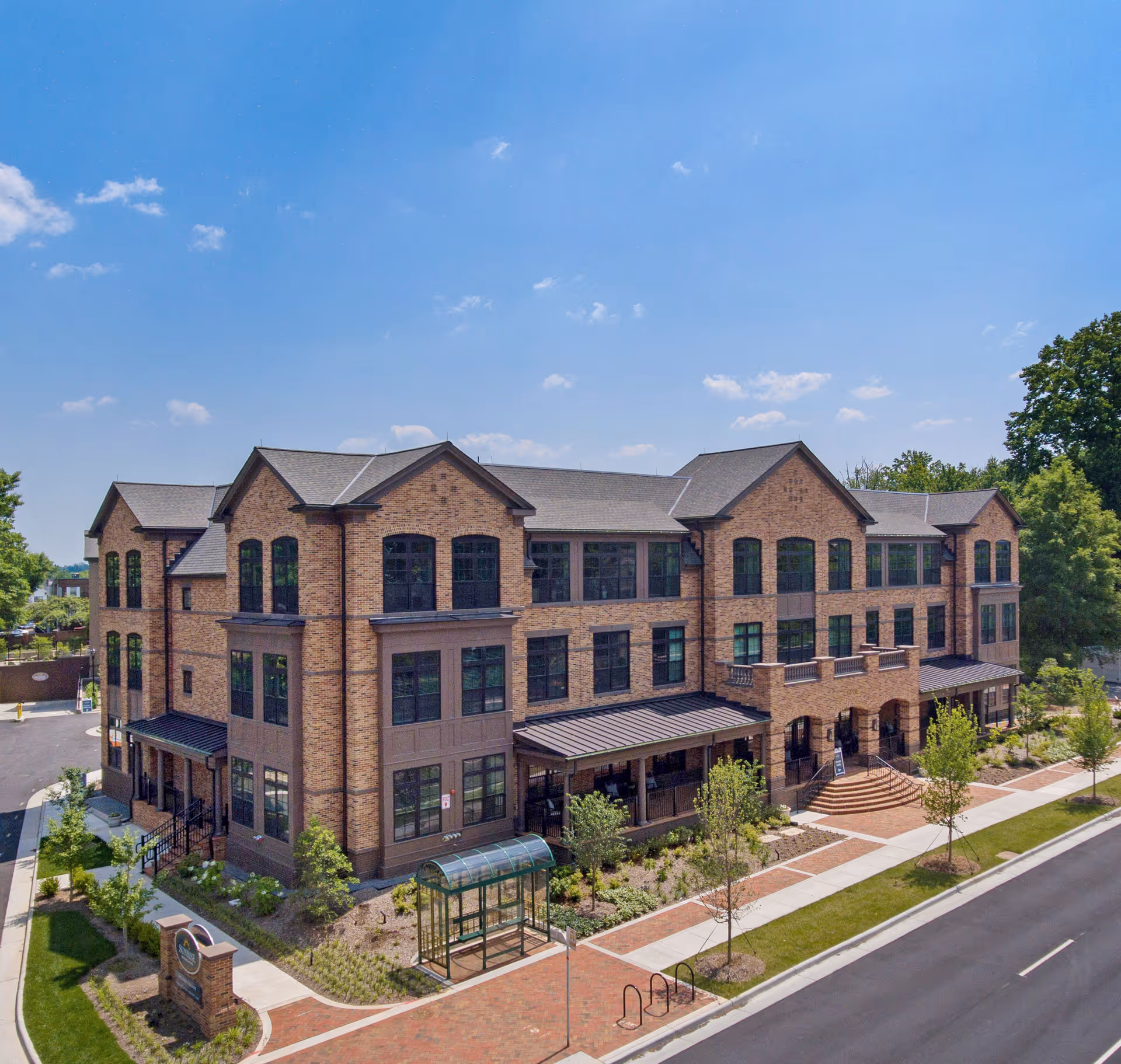 Three-story brick senior living building with a landscaped entrance, sidewalks and a bus shelter under a clear blue sky.