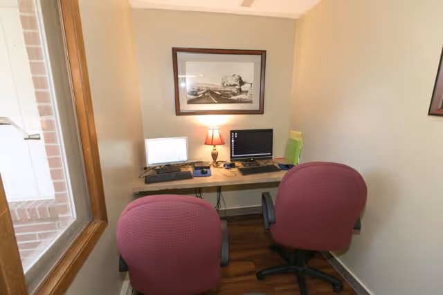 Small office space with two red office chairs facing a wall-mounted desk holding two computer monitors, a keyboard, a mouse, a small lamp, and some office supplies. A framed black and white photograph hangs on the wall above the desk. There is a window on the left side showing a brick exterior wall.