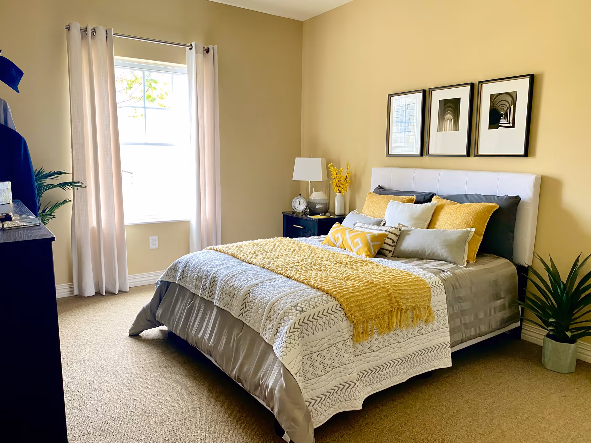 A neatly made bed with a white and gray patterned comforter, yellow throw blanket, and multiple pillows in shades of yellow, gray, and white. The bed has a white upholstered headboard. On the right side of the bed is a black nightstand with a lamp, clock, and a vase with yellow flowers. Above the bed are three framed black and white photographs. A window with white curtains is on the left wall, and there is a green potted plant on the floor near the bed. The walls are painted a soft yellow color and the floor is carpeted.