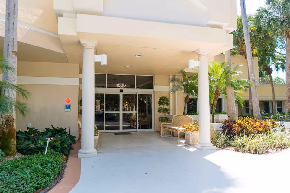 Front covered entrance of a senior living building with columns, glass double doors marked "1600", a bench and surrounding landscaping.