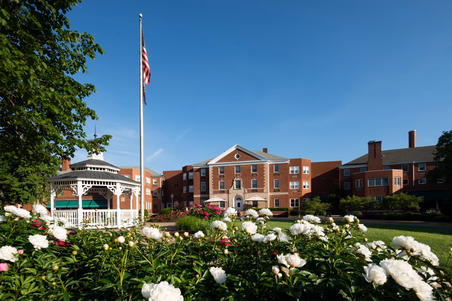 Front lawn and entrance of a red-brick senior living building with a white gazebo, flagpole, and blooming white flowers under a clear blue sky.