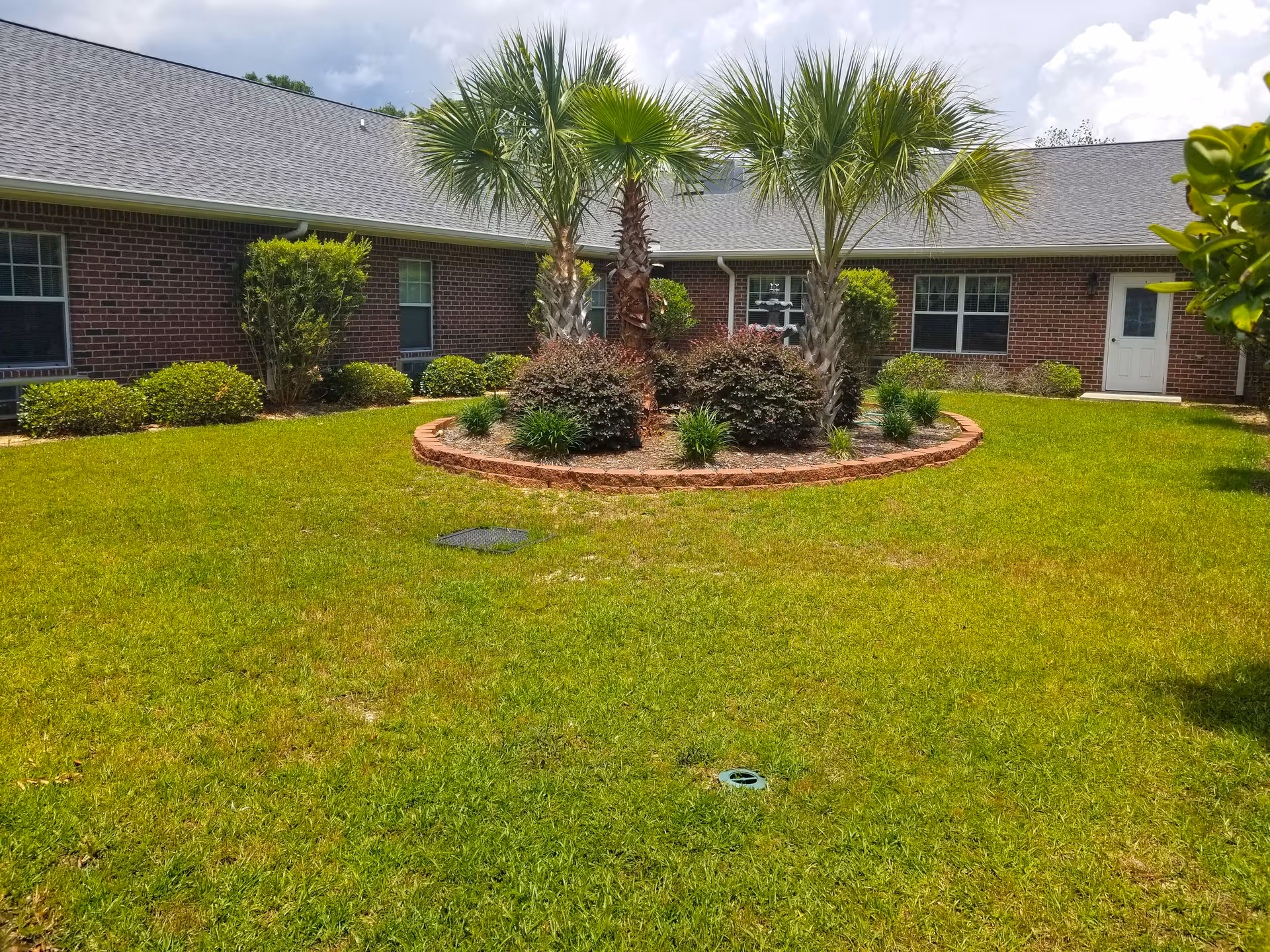 A courtyard garden area with green grass and a circular flower bed containing palm trees and shrubs, surrounded by a single-story brick building with windows and a white door under a partly cloudy sky.
