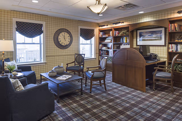 A cozy senior living common area with plaid carpet and wallpaper. The room features a seating area with a sofa, armchairs, and a coffee table with decorative items. There are two windows with patterned valances, a large wall clock, and built-in wooden bookshelves filled with books and decor. A wooden desk with a computer and chair is situated near the bookshelves. The lighting includes a ceiling fixture and a table lamp.