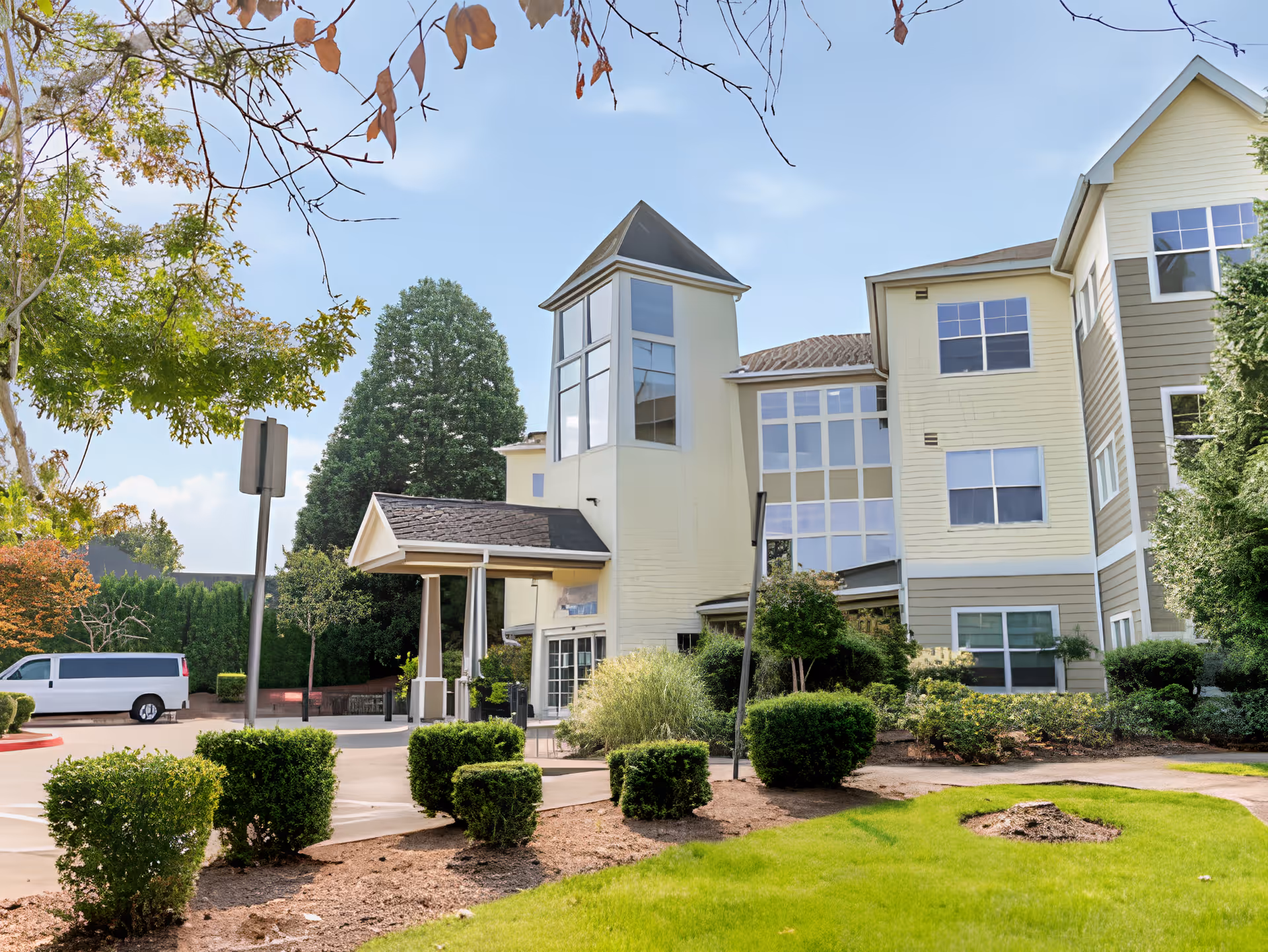 Exterior view of a multi-story senior living facility building with large windows and a covered entrance. The building is surrounded by neatly trimmed bushes, trees, and a well-maintained lawn under a clear blue sky.