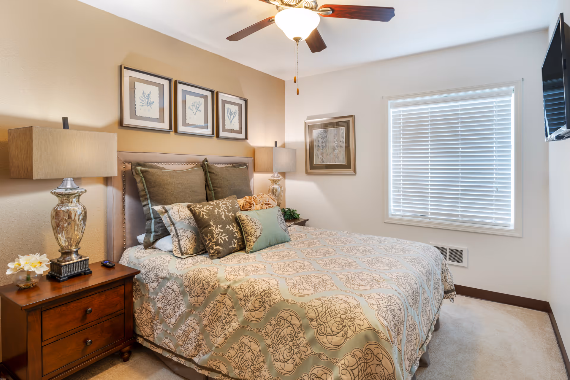 A neatly made bed with patterned bedding and multiple decorative pillows in a bedroom. There are two bedside tables with matching lamps, framed artwork on the walls, a ceiling fan with light, a window with blinds, and a wall-mounted TV.