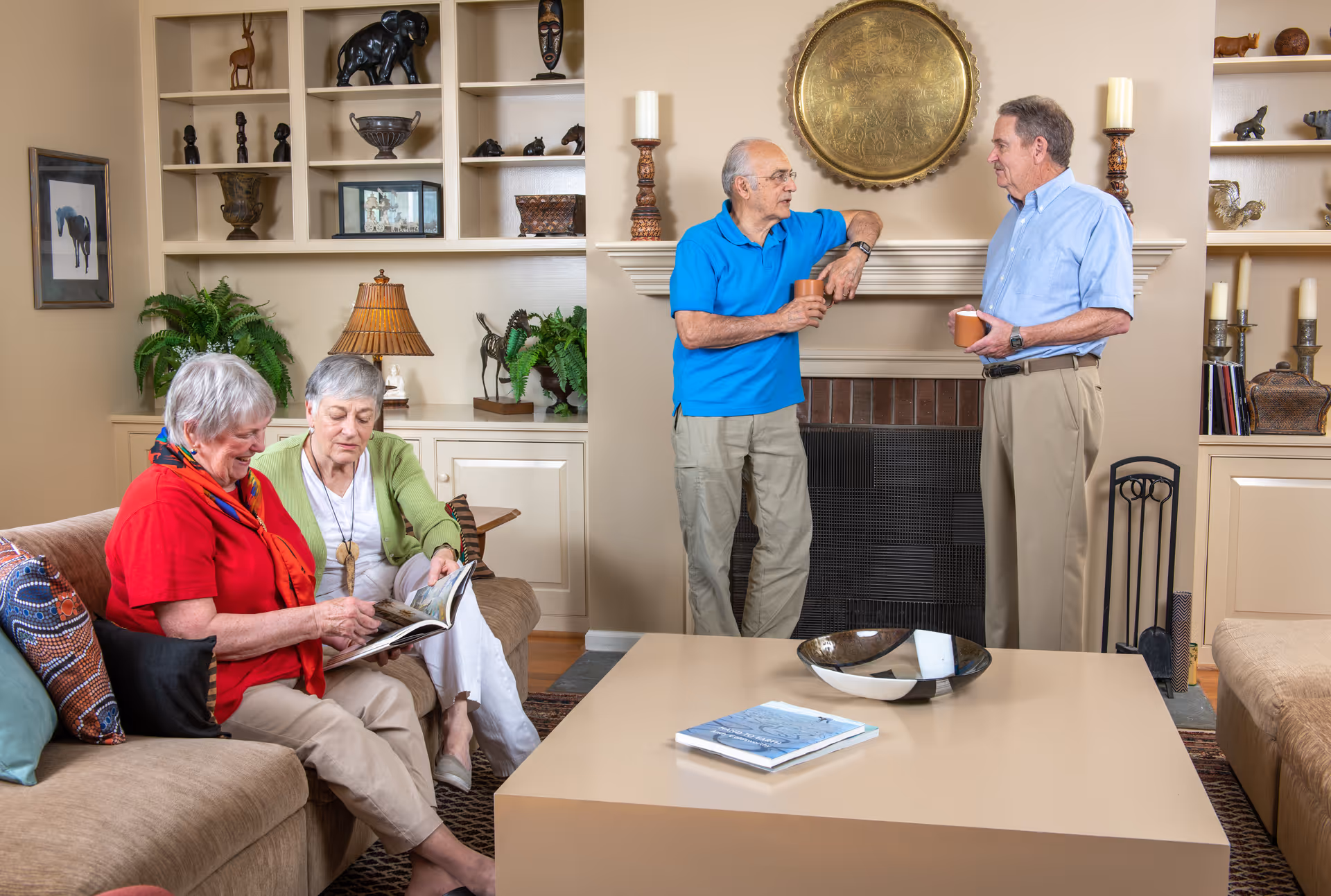 Two elderly women sitting on a beige couch looking at a book together, while two elderly men stand near a fireplace holding mugs and talking in a cozy living room with built-in shelves, decorative items, and plants.