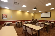 A dining room with several rectangular tables and chairs arranged neatly. The walls are painted beige with a wooden trim, and there is a large white projection screen on one wall. The ceiling has recessed lighting and a projector mounted in the center.