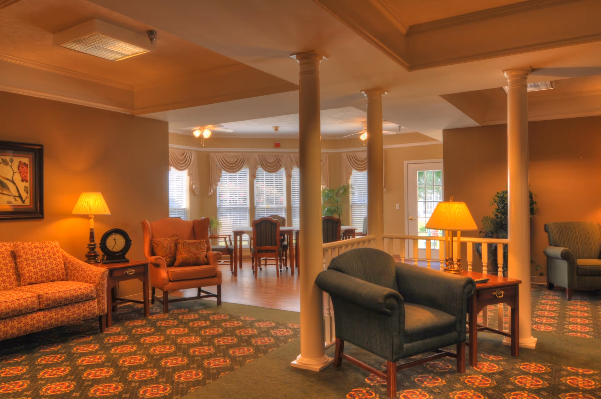 A cozy senior living facility common area with comfortable armchairs and sofas arranged around small wooden side tables with lamps. The room features decorative columns, patterned carpet, and large windows with draped curtains letting in natural light. A dining area with wooden chairs and tables is visible in the background.