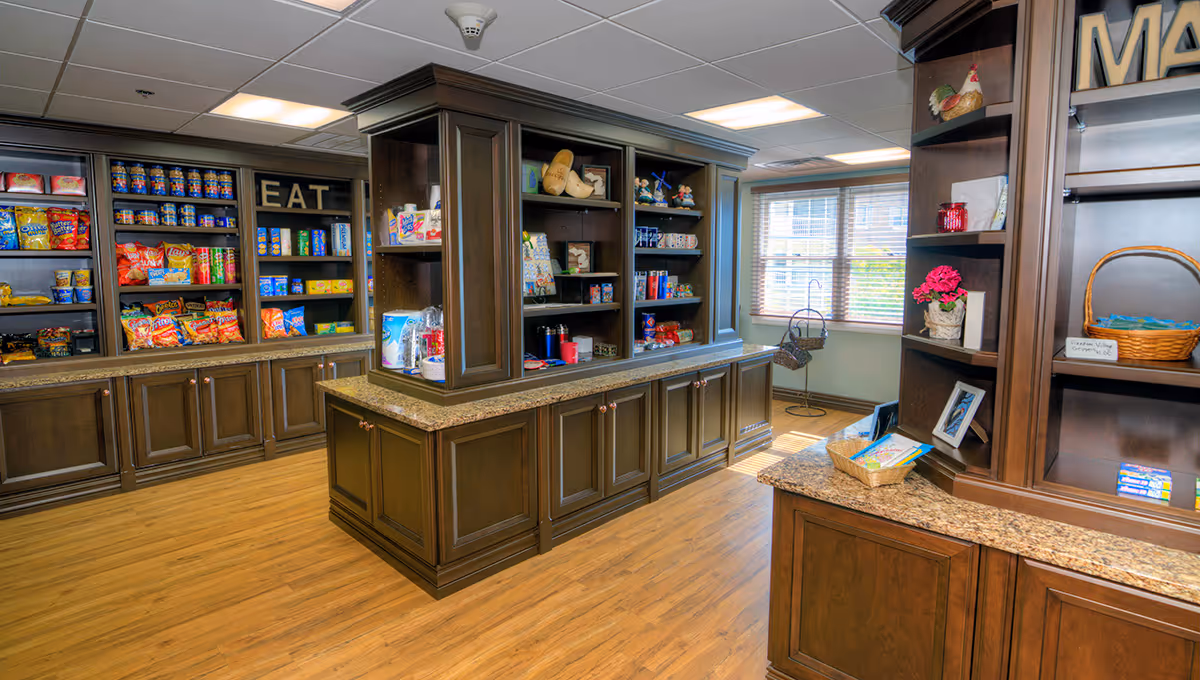 Interior view of a snack and convenience area in a senior living facility with wooden shelves and cabinets stocked with various snacks and food items. The room has wood flooring, a window with blinds, and decorative items on the shelves.