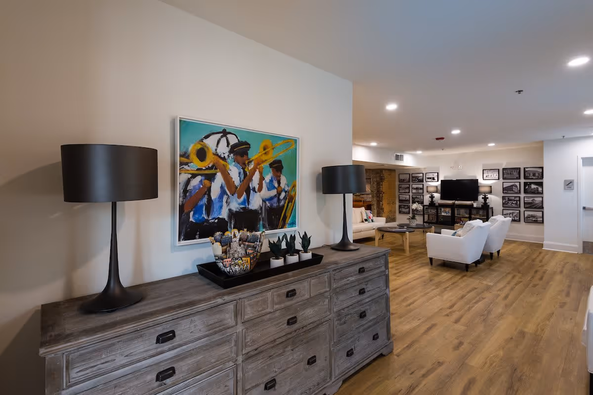 A spacious and well-lit living room area with wooden flooring. In the foreground, there is a rustic wooden dresser with two black lamps, a tray with small plants, and decorative items. Above the dresser hangs a colorful painting of musicians playing trombones. In the background, there are white armchairs, a coffee table, a TV mounted on a stand, and walls adorned with multiple framed black and white photographs.