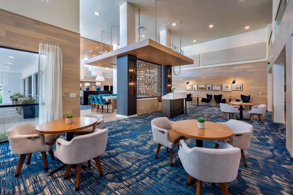A spacious and modern common area with round wooden tables and cushioned chairs arranged on a blue patterned carpet. The room features light wood-paneled walls, framed artwork, and a central decorative partition with the word 'OVATION' above it. Large windows with sheer curtains allow natural light to enter, and there is a view of an outdoor patio area.