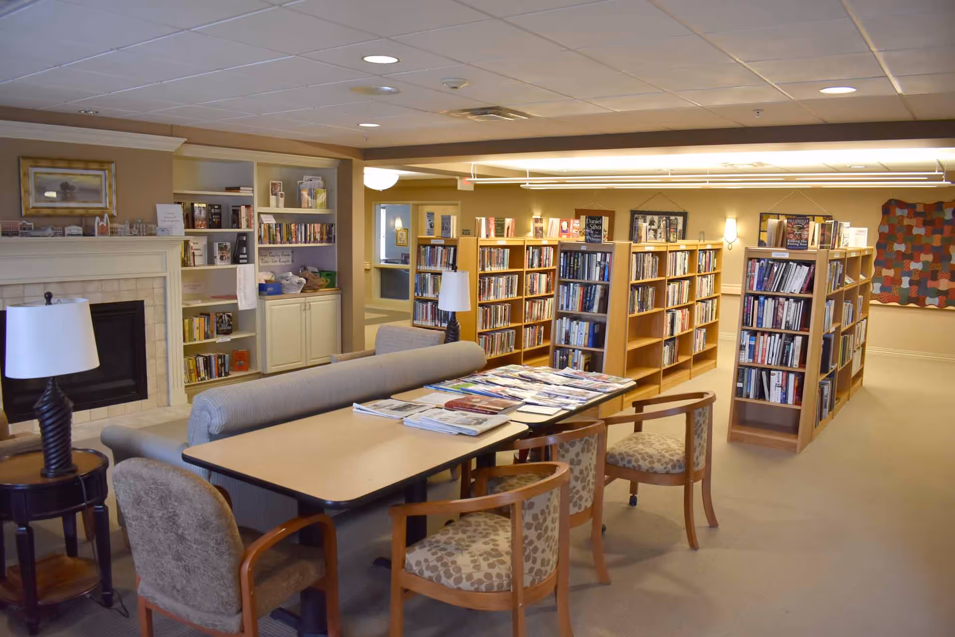A cozy library or reading room with bookshelves filled with books, a table with chairs, a couch, two table lamps, and a fireplace. The room has a warm, inviting atmosphere with soft lighting and a quilt hanging on the wall.